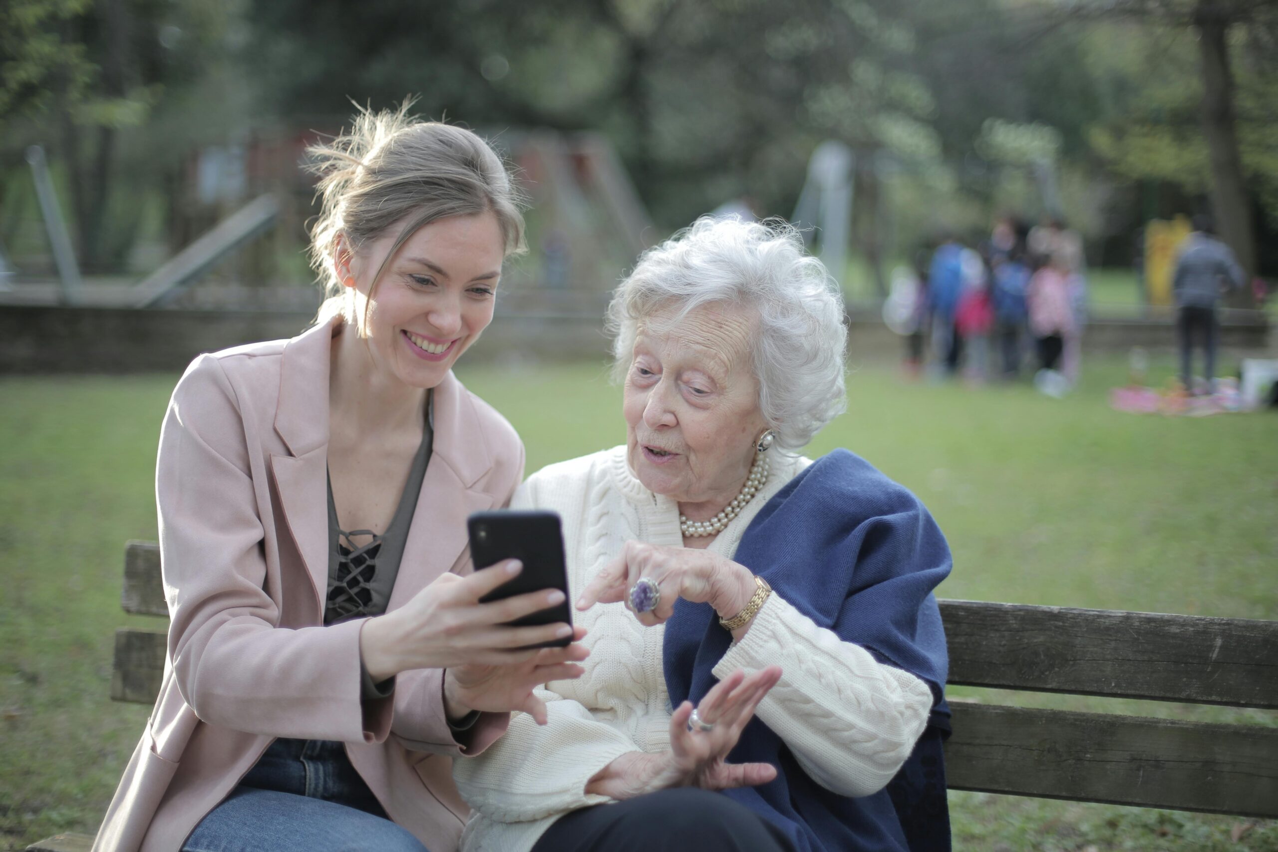 Female relatives sitting together on wooden bench in park, browsing mobile phone to learn how to use Telegramsy services