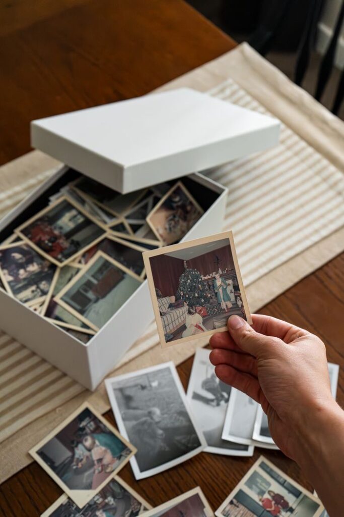 pexels-photo-34384409-34384409 A hand holding a vintage family photo above a box of nostalgic family snapshots on a table.